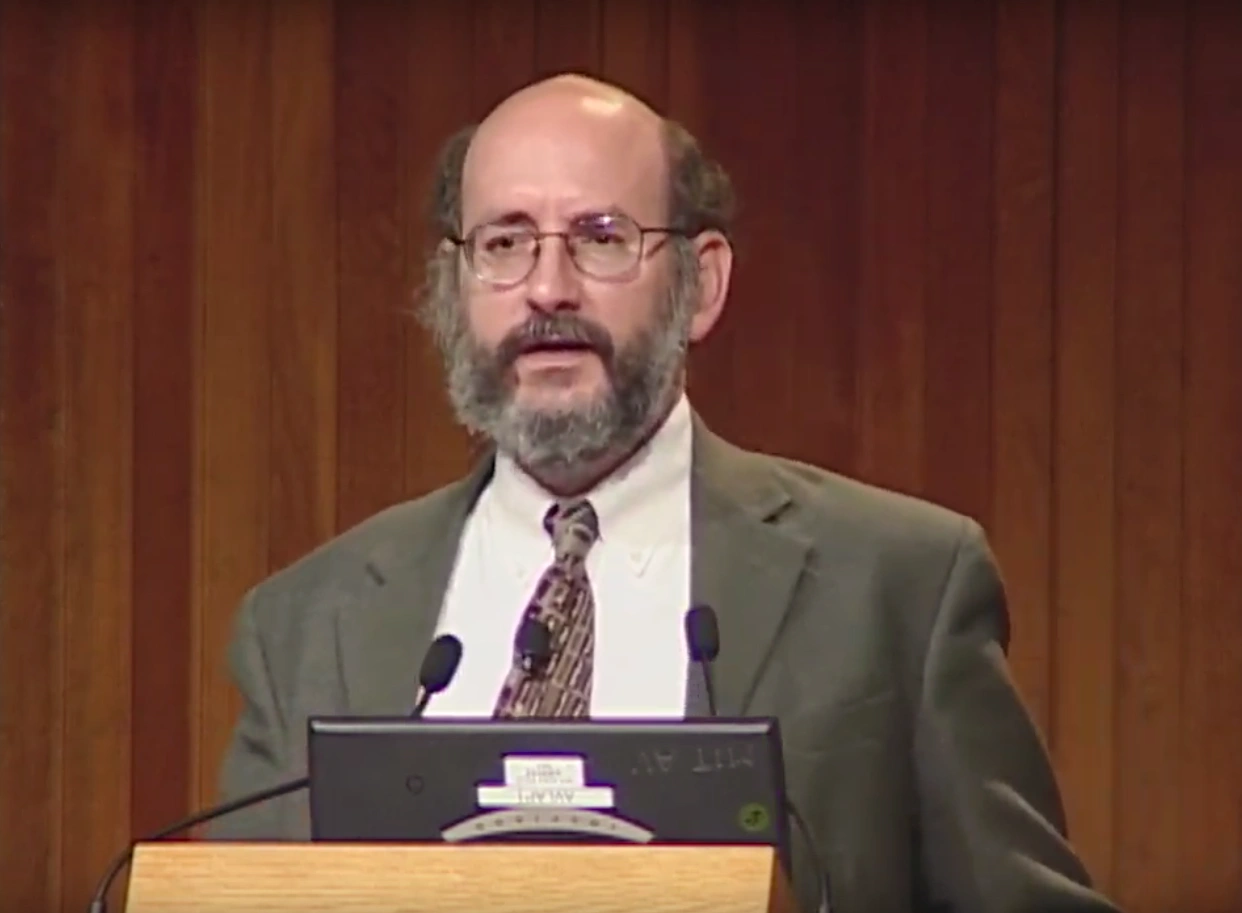Larry Vale speaks onstage, standing behind a podium, red curtain in the background