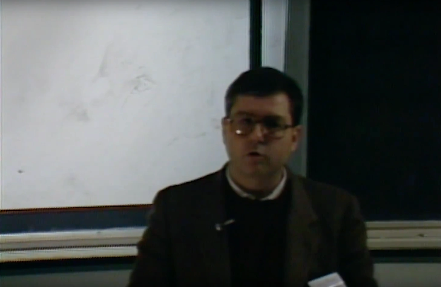 A man stands with arms crossed in a classroom, projector screen and chalkboard in background