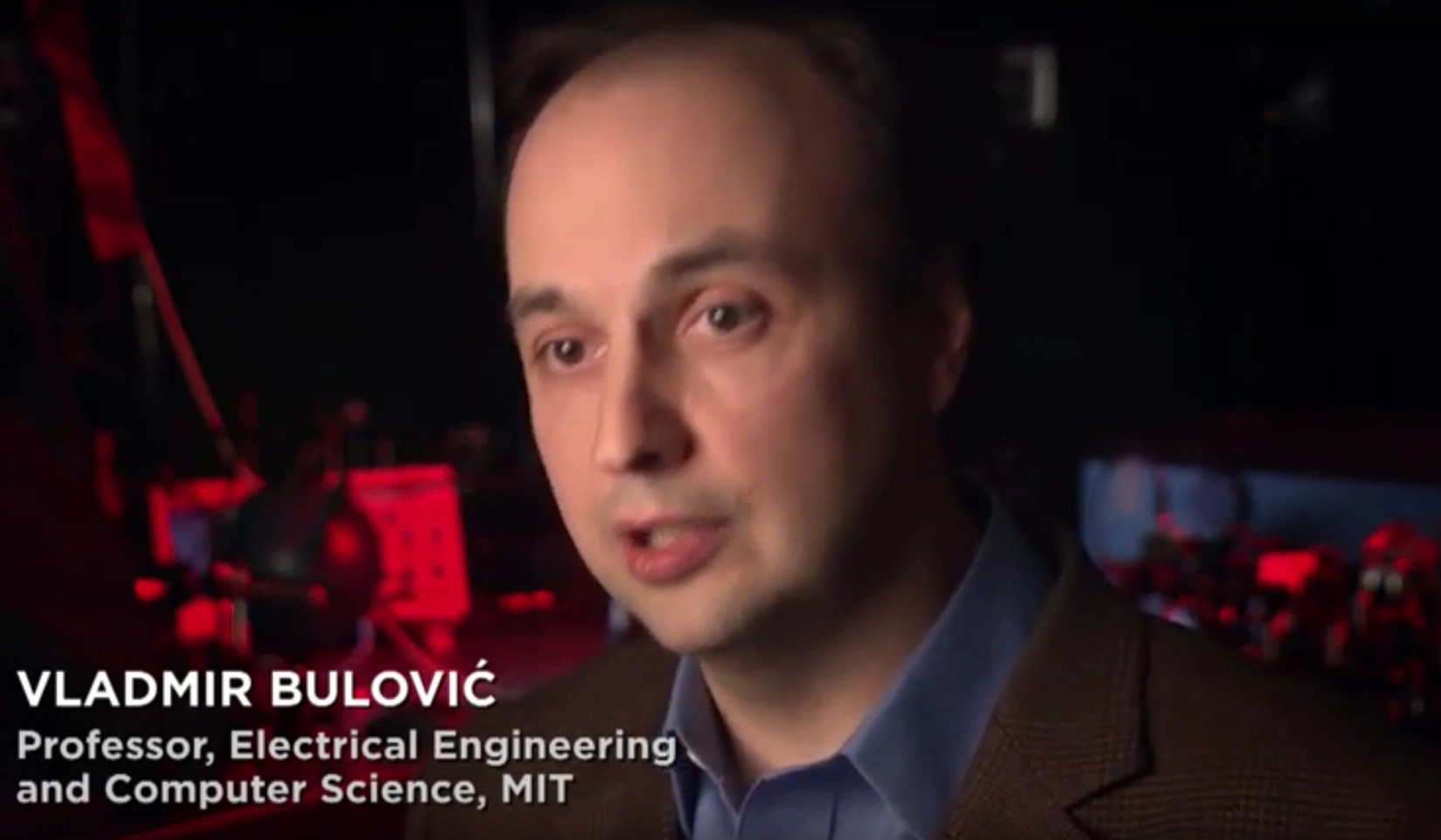 Vladmir Bulović mid-sentence during seated on camera interview, dim red light illuminates computers in the background.