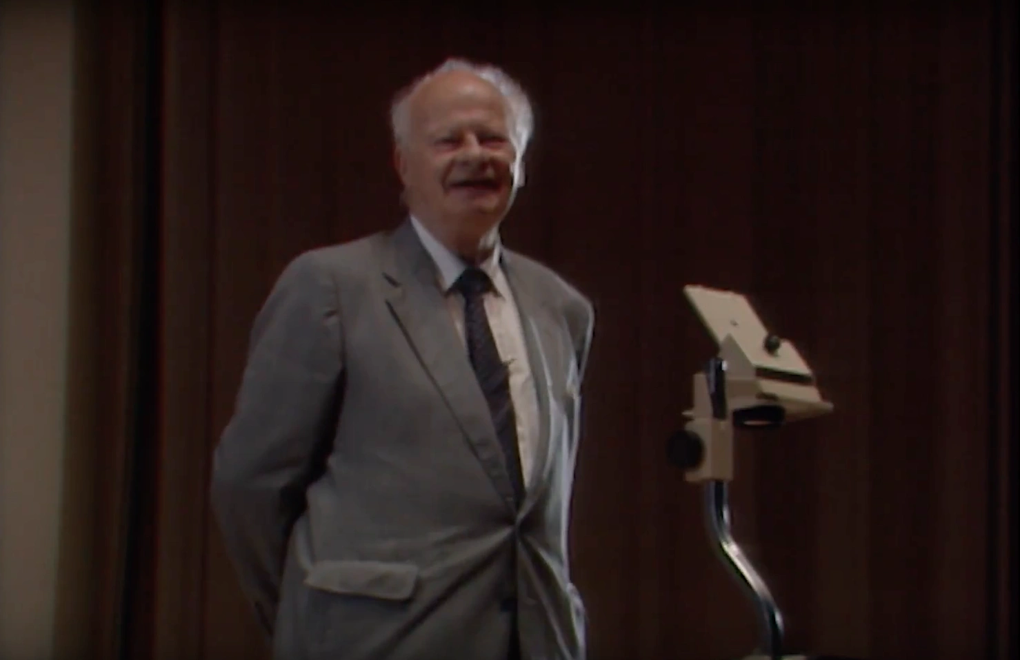 Hans A. Bethe standing beside optical microscope on dimly lit stage, cream colored curtain in background.