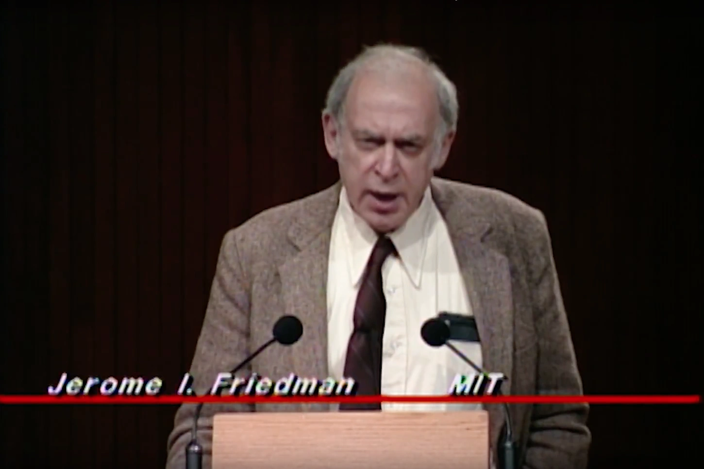 Jerome I. Friedman speaking at a podium on stage, in front of a dark background