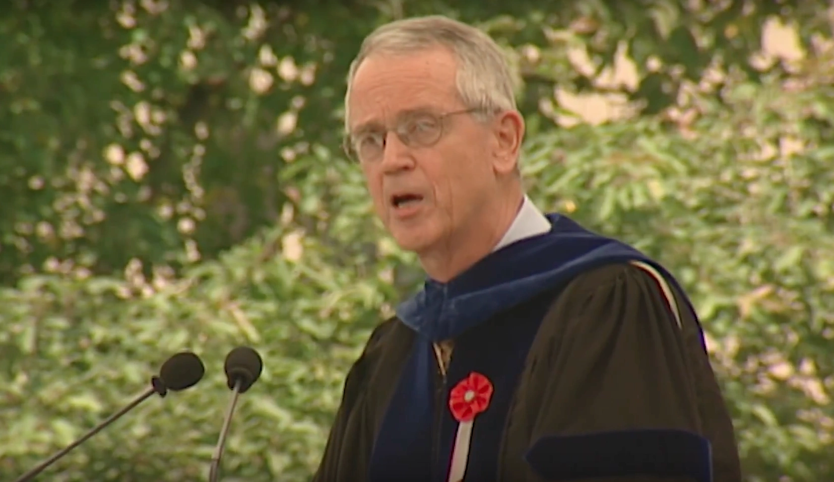 Charles M. Vest, wearing commencement attire, speaking at podium on outdoor stage, trees in background