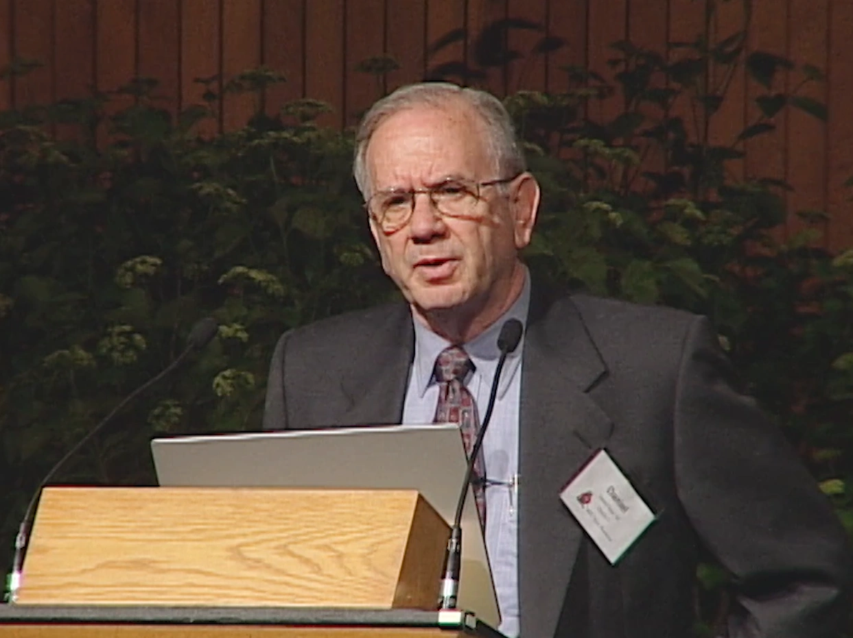 Mike Fincke speaks at podium onstage, leaves from a potted plant and red curtain in background