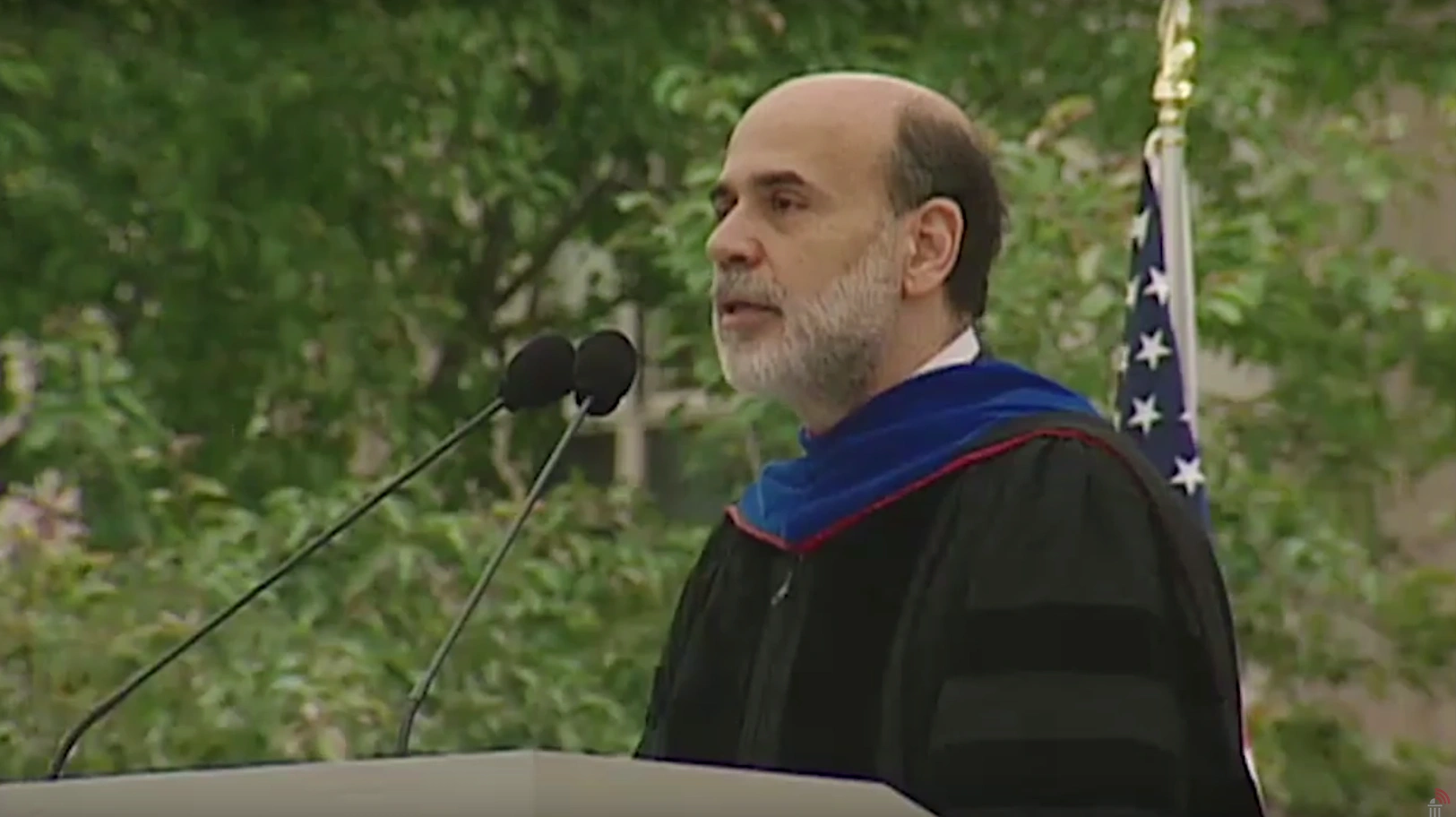 Ben Bernanke, wearing commencement attire, speaking at podium on outdoor stage, trees in background