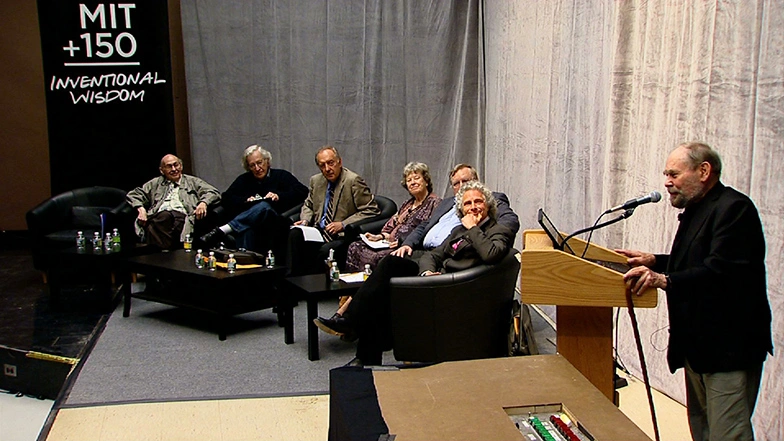 Group of panelists seated chairs on stage, watching a man speak standing behind a podium, light gray curtains in background.