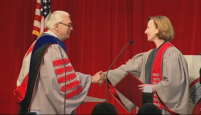 Paul E. Gray and Susan Hockfield, both wearing commencement attire, shake hands on stage, red curtain in background.