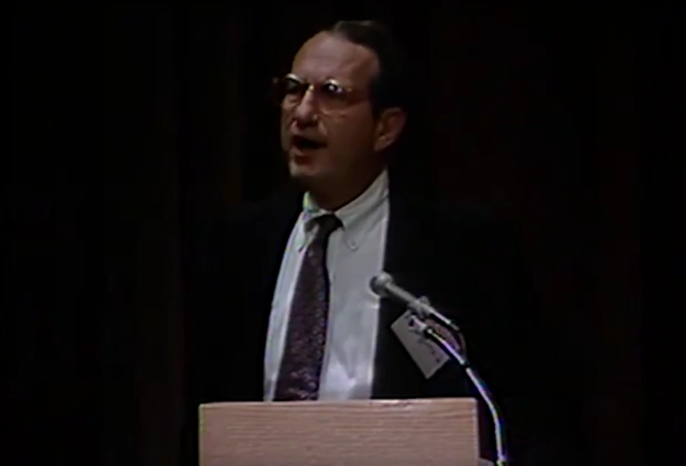 John M. Deutch speaking at a podium on stage, in front of a dark background