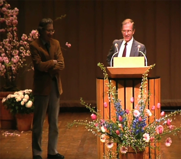 Noam Chomsky speaks on stage, a large arrangement of flowers placed in front of his podium and a red curtain in the background.