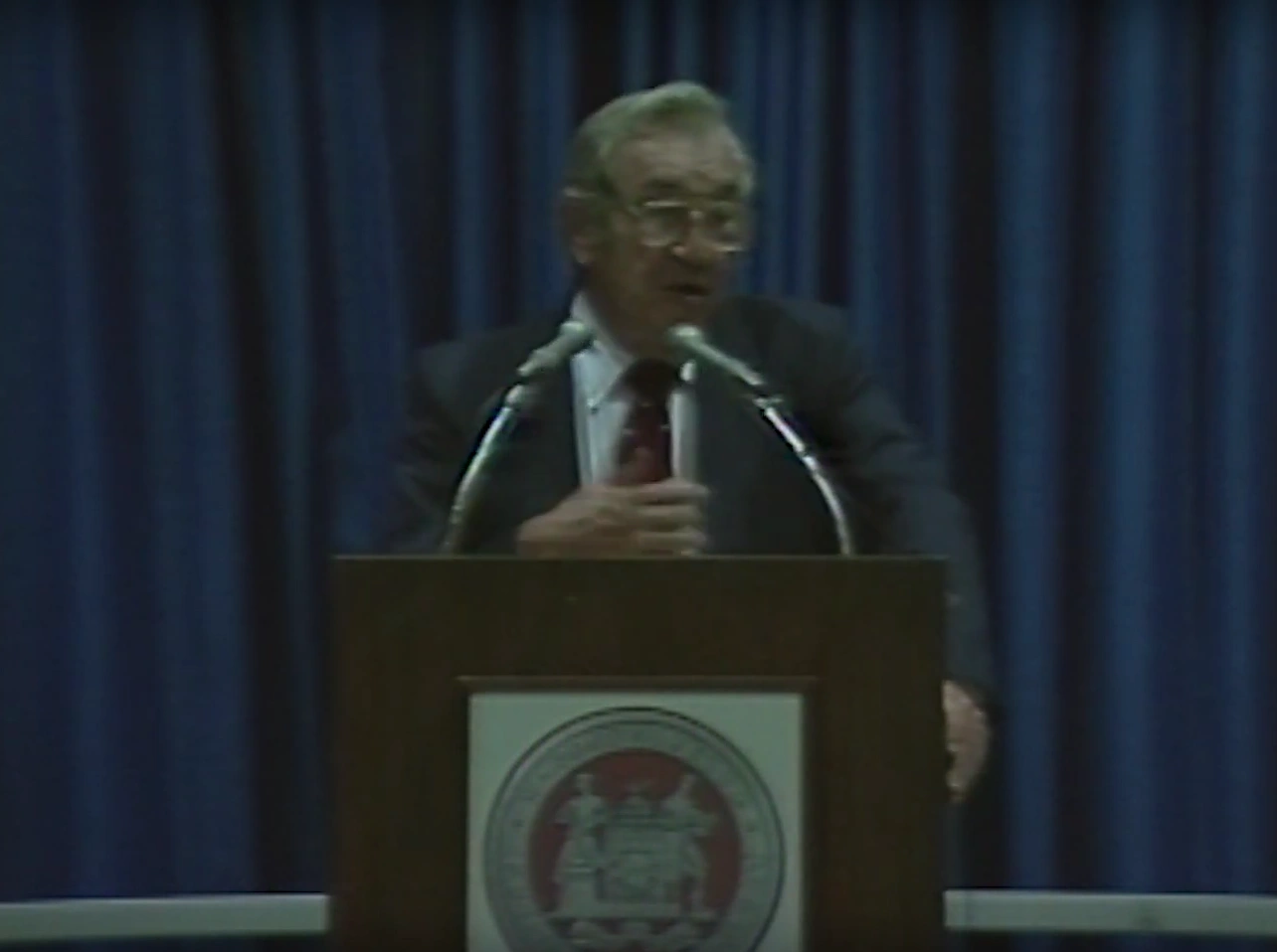 Jerome Wiesner stands behind a podium microphone, looking ahead. A marble wall in the background.