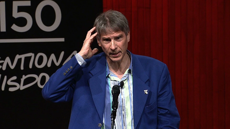 Christof Koch speaking at a podium onstage, pressing his fingertips to one side of his head, red curtain in background.
