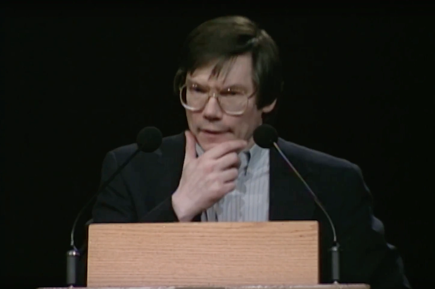 Alan Guth standing on stage at podium, hand resting on chin in thinking pose, dark background
