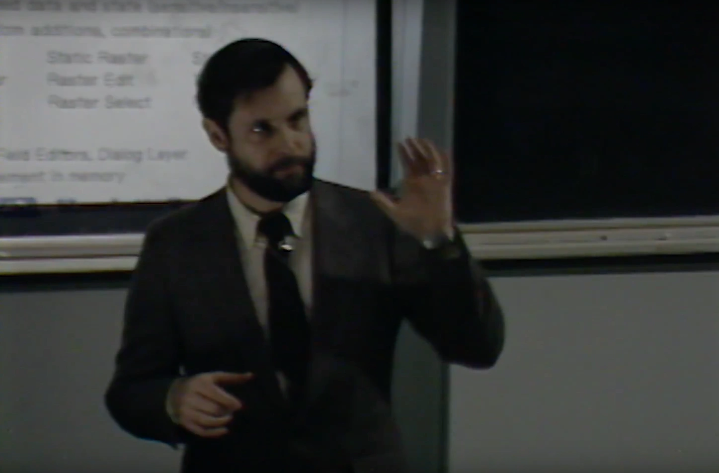 A man lectures at a podium in a classroom, projector screen and chalkboard in background