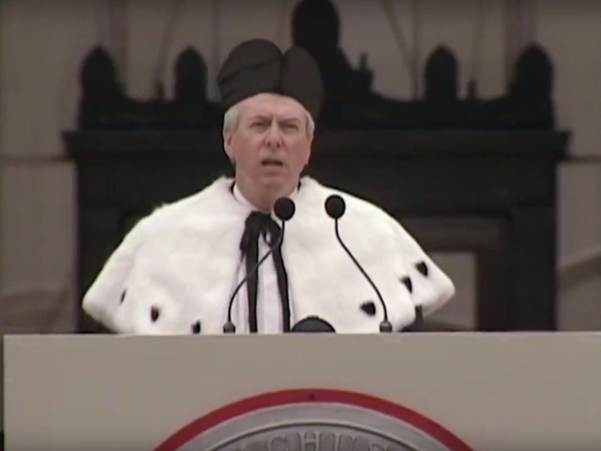 Daniel S. Golden, wearing commencement attire, speaking at podium on outdoor stage, trees in background