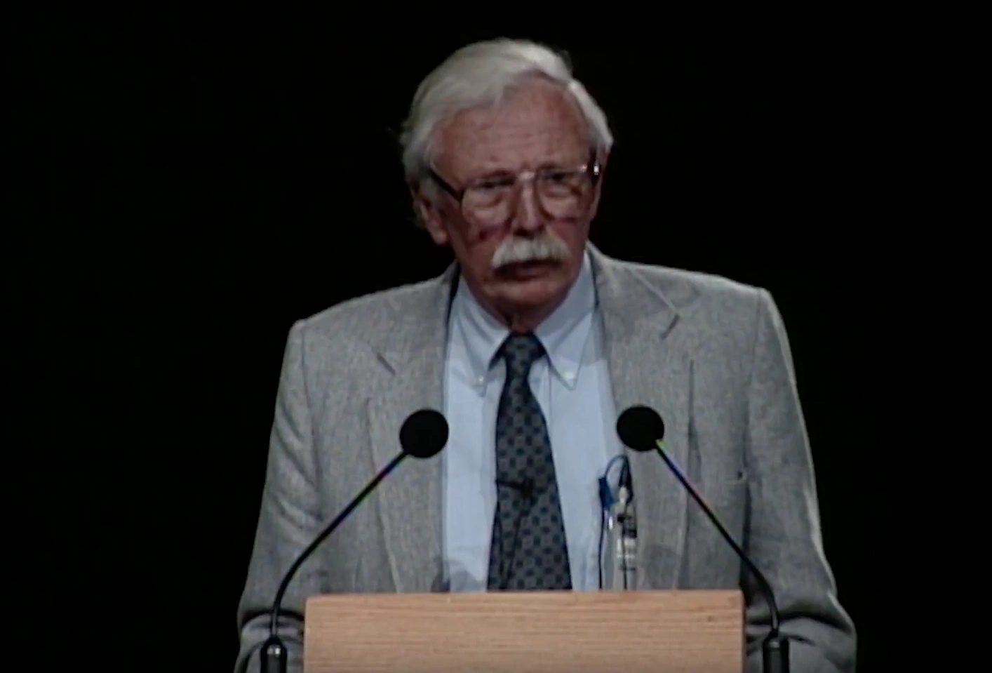 William L. Kraushaar speaking at a podium on stage, in front of a dark background