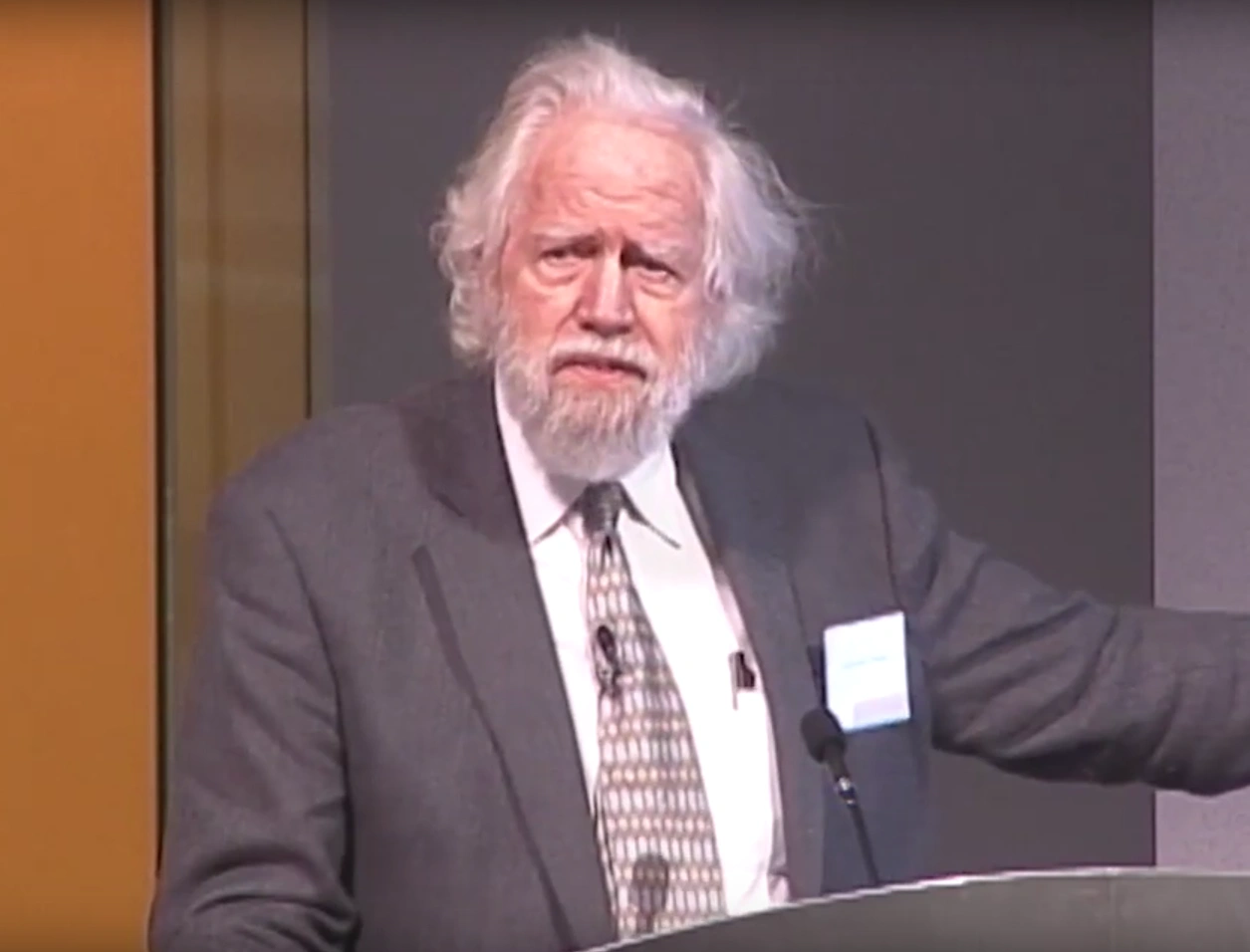 Alexander Shulgin speaks at a podium infront of a gray background