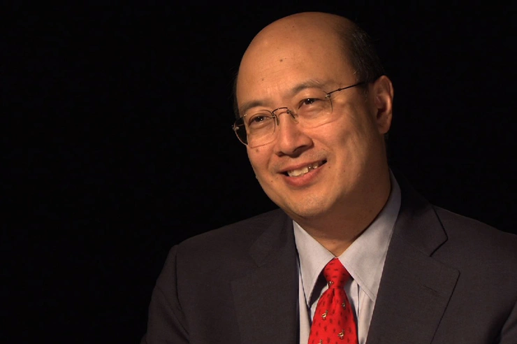 Andrew Lo seated in front of a black background for an on camera interview, smiling.