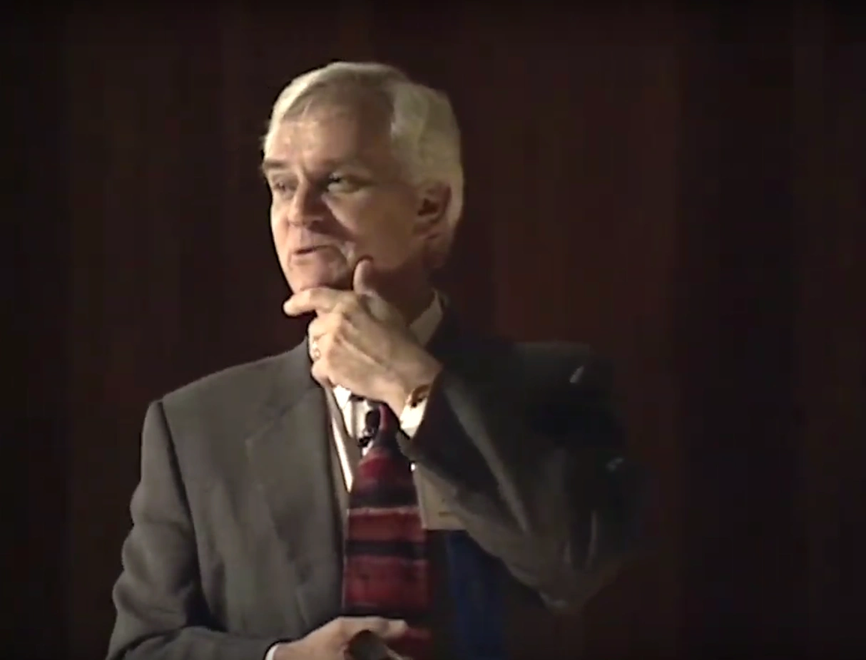 Charles M. Vest stands on stage, hand resting on chin in thinking pose, black curtain in background.