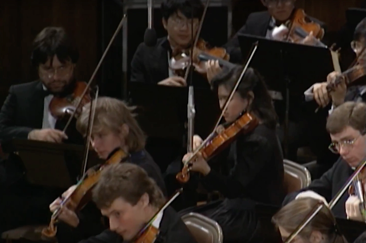 Violin section of orchestra, dressed in matching black formal wear, stare at sheet music while playing intently.