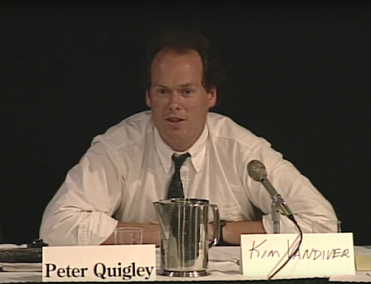 Peter Quigley speaks into podium microphone, seated behind his name plate at a table