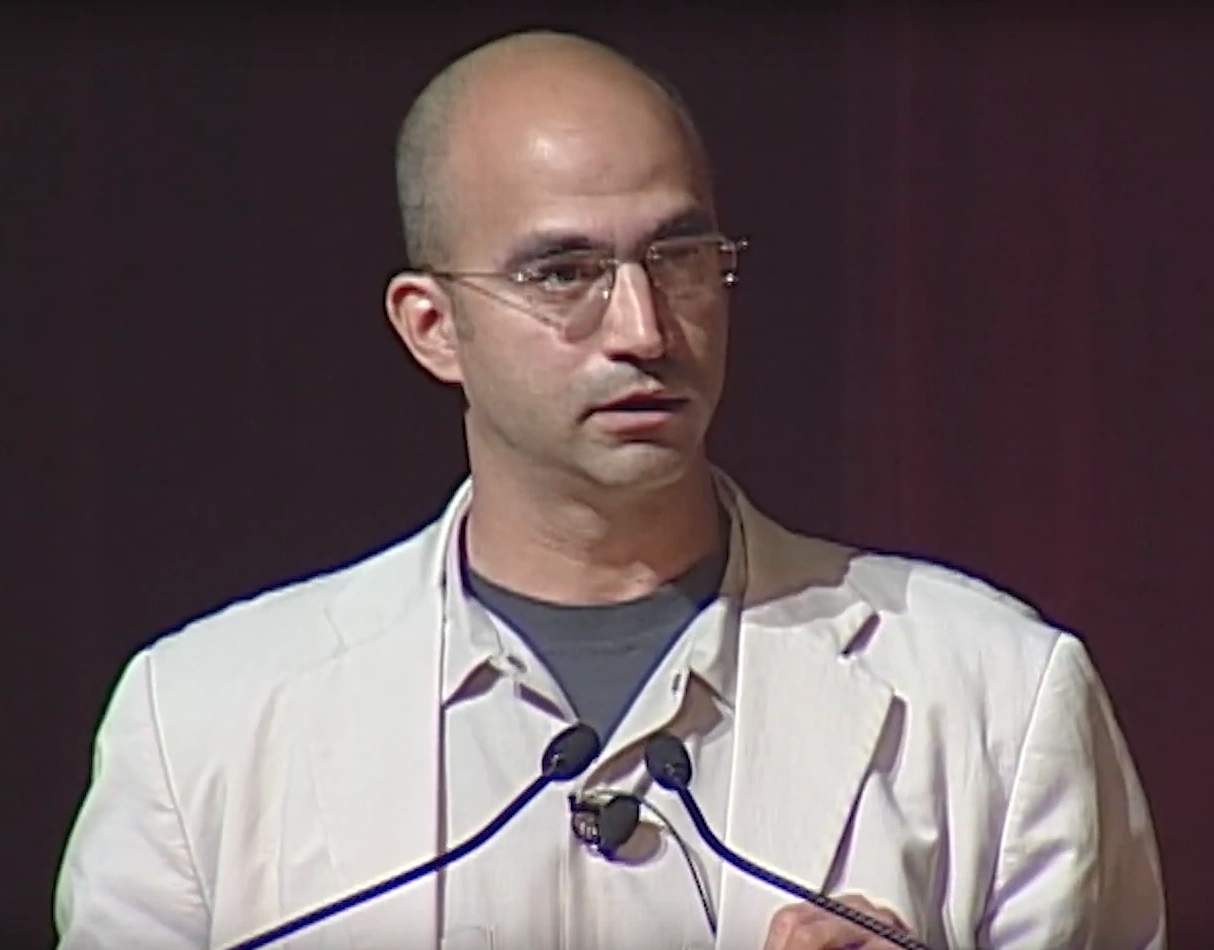 John Fernandez speaking into a podium microphones onstage, dark curtian in the back. 
