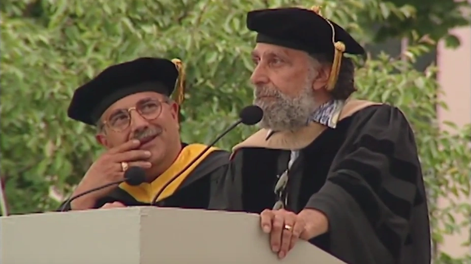 Ray and Tom Magliozzi dressed in commencement attire speaking at a podium outside in front of trees