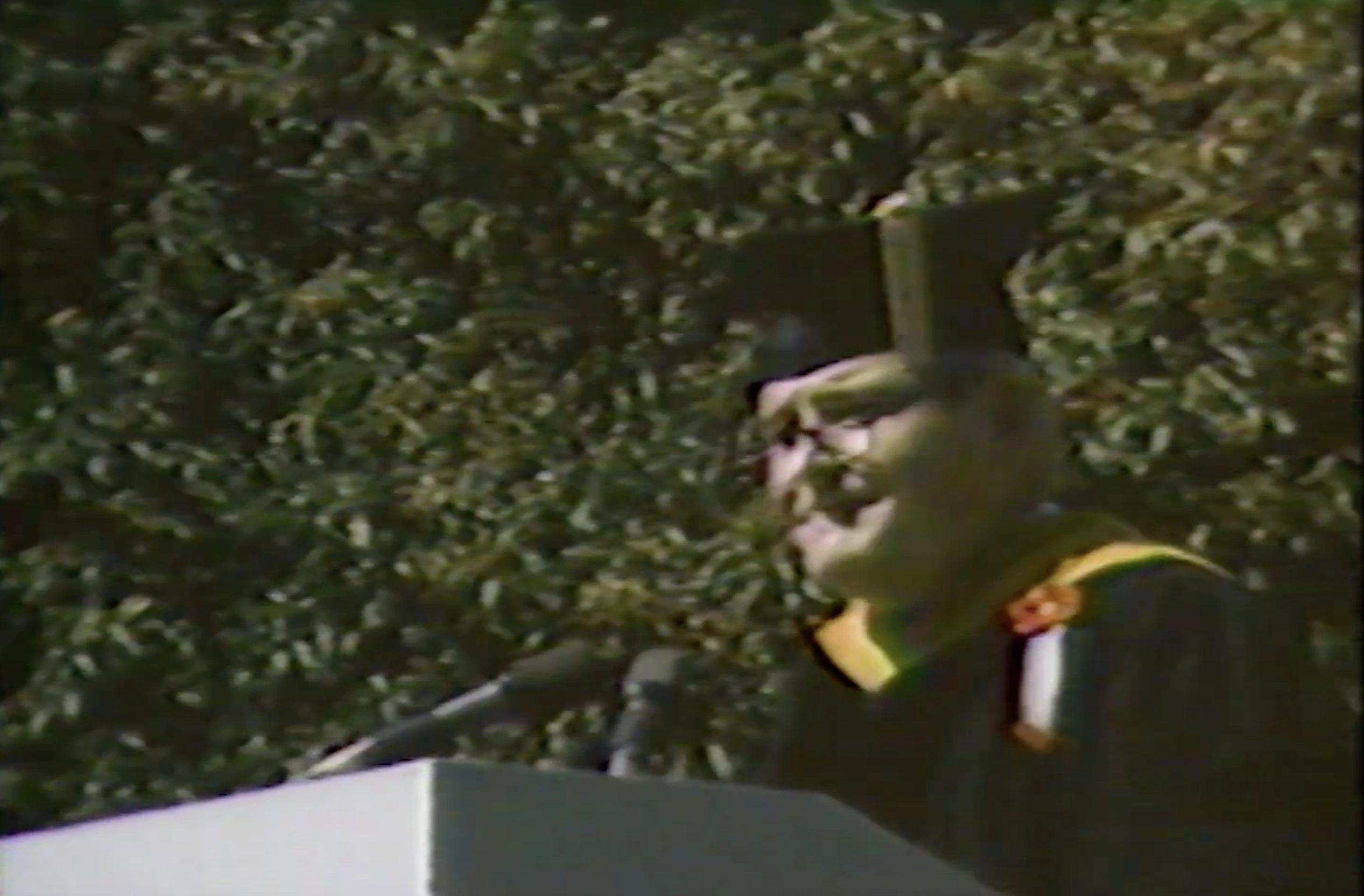 Paul E. Gray, wearing commencement attire, speaking at podium on outdoor stage, trees in background