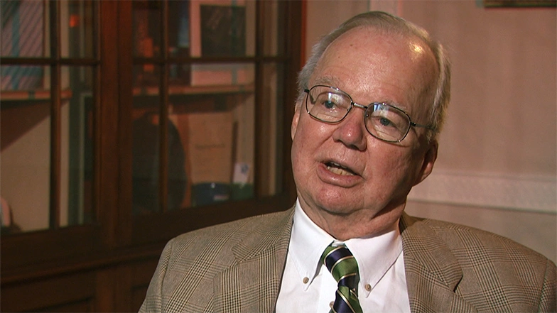 Howard Johnson seated in room for on camera interview, mid-sentence. White walls in background