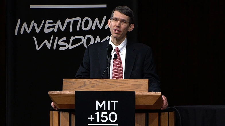 James M. Poterba speaking at a podium on stage, in front of a dark background
