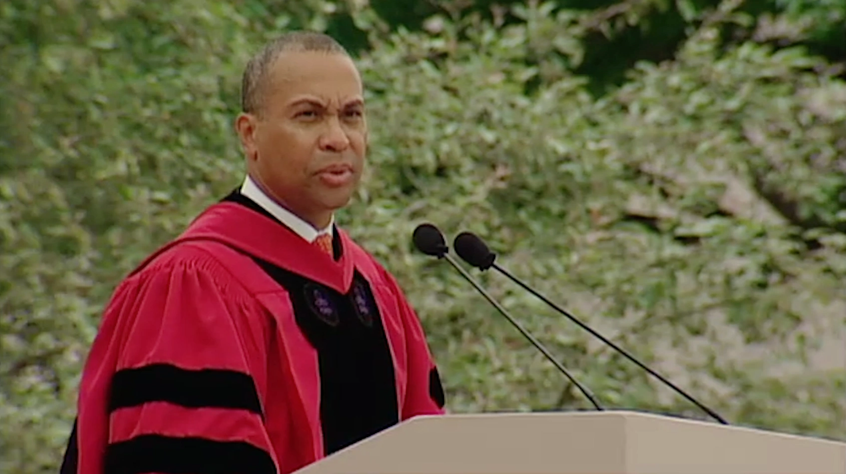 Deval Patrick, wearing commencement attire, speaking at podium on outdoor stage, trees in background