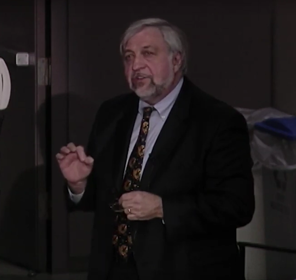Jerry Melillo delivers speech in dimly lit room in front of a chalkboard