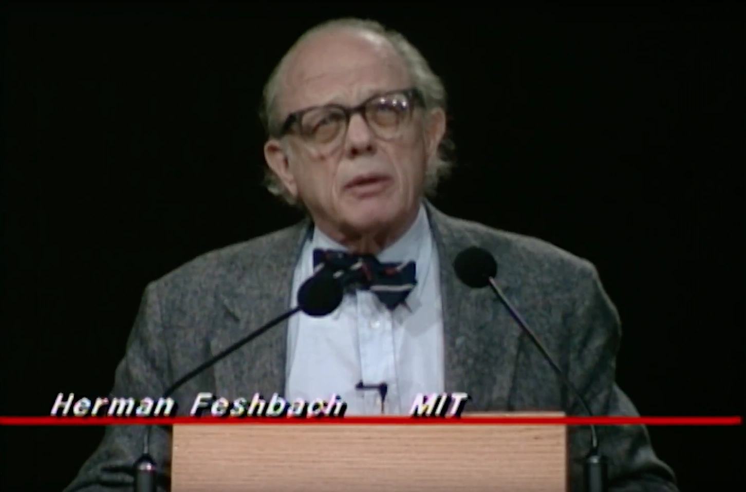Herman Feshbach speaking at a podium on stage, in front of a dark background