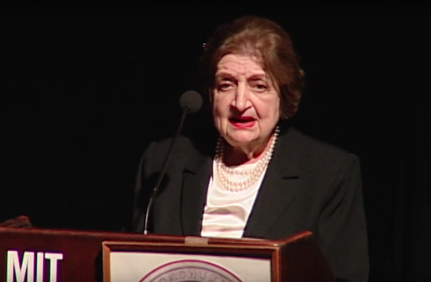Helen Thomas speaks at a podium onstage in front of black curtain