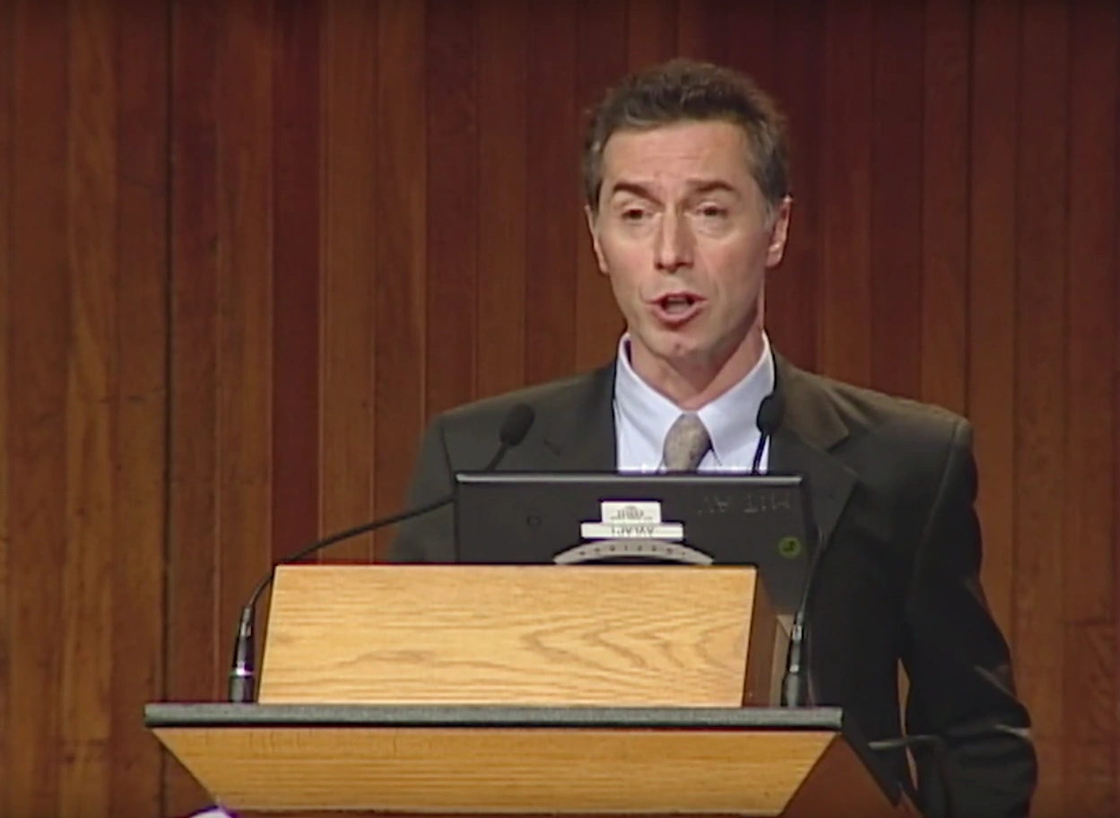 Richard Locke speaks onstage, standing behind a podium, red curtain in the background