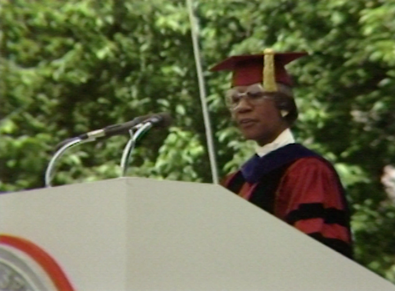 Shirley Chisholm, wearing commencement attire, speaking at podium on outdoor stage, trees in background