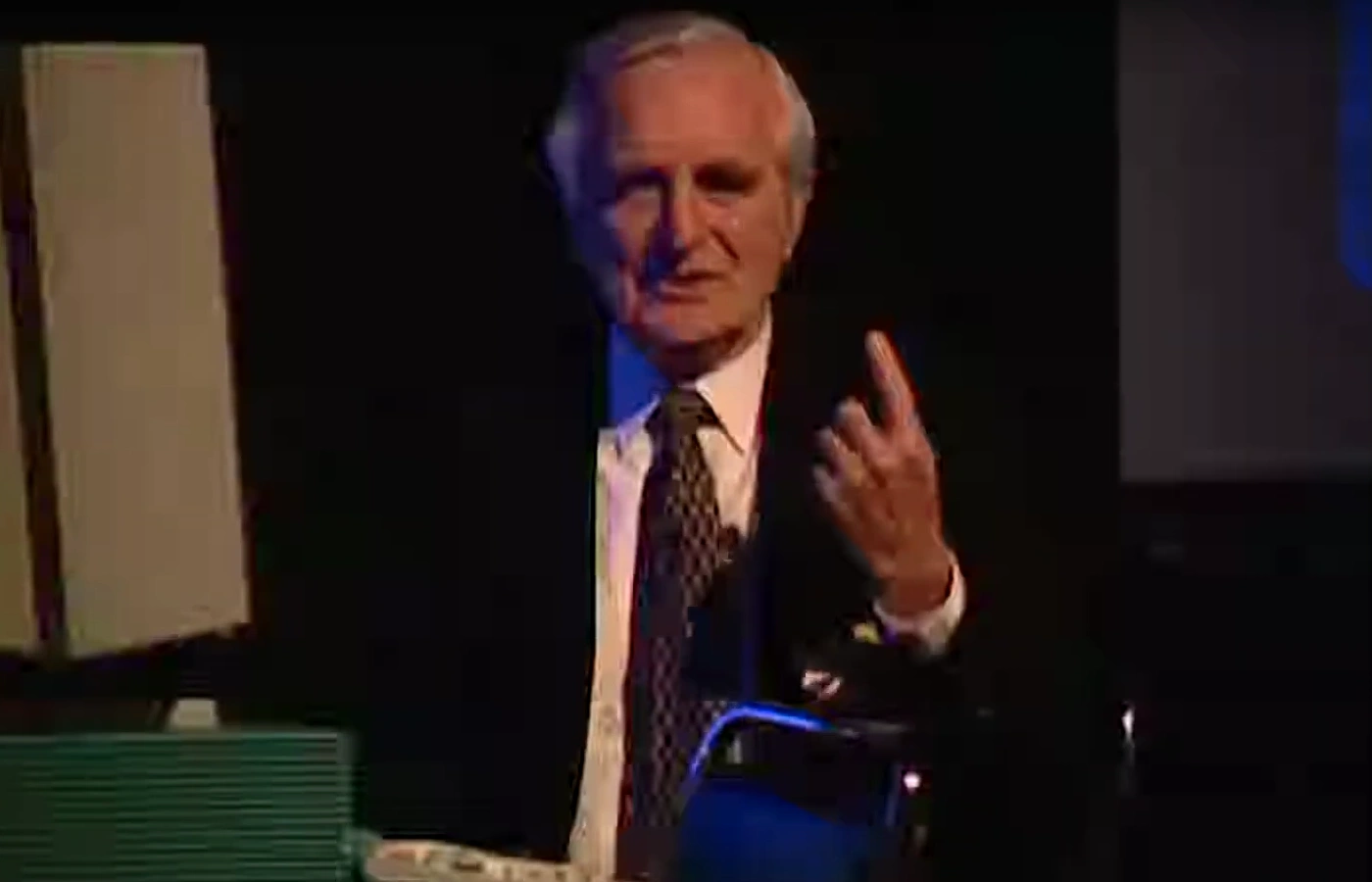 Paul Penfield holding up his index finger to gesture while speaking on dimly lit stage