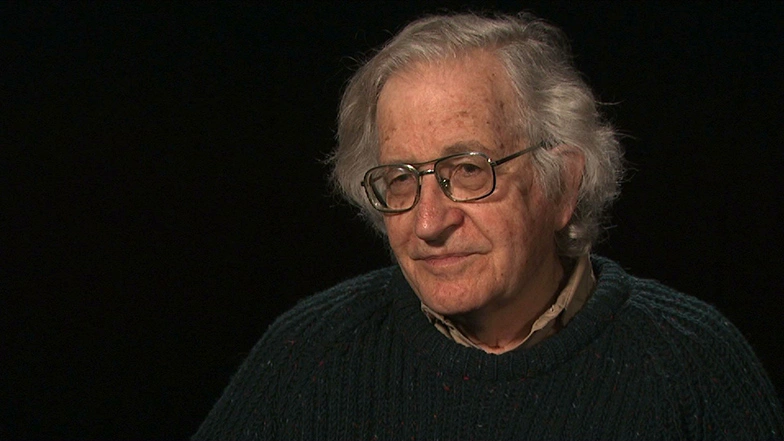 Noam Chomsky seated in front of a black background for an on camera interview, smiling.