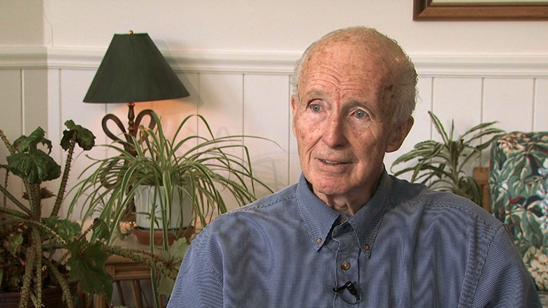Joseph G. Gavin, Jr. seated in white room with ficus plants in the background, looking ahead during an on camera interview.