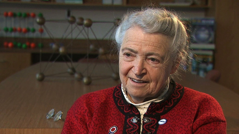 Mildred Dresselhaus seated and smiling for an on camera interview, models of molecules in background