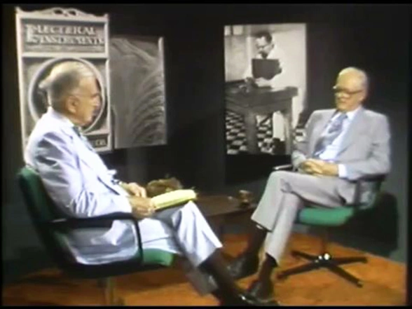 A conversation between Lauriston S. Taylor and John G. Trump on stage, large framed posters behind them