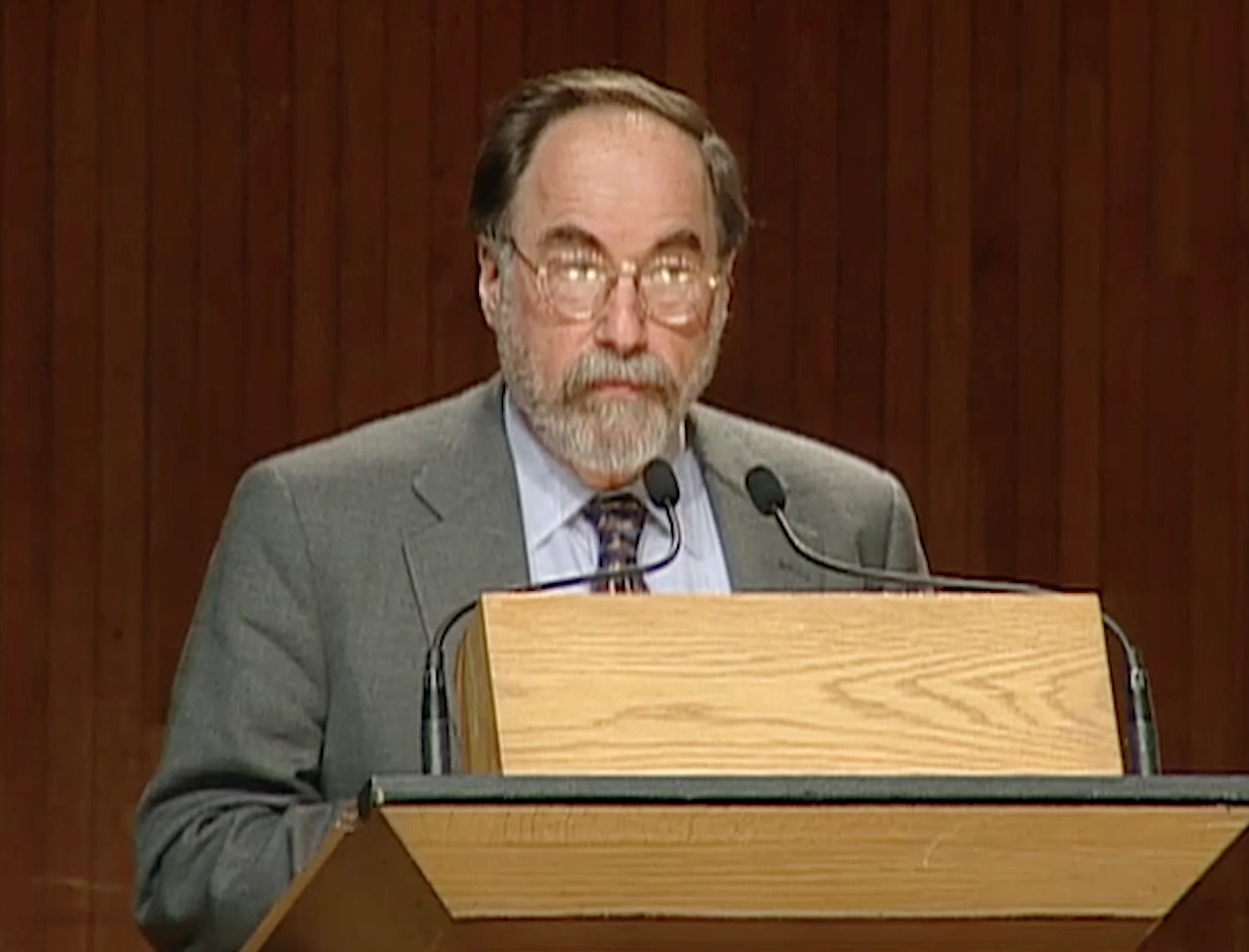 David Baltimore standing at a podium on stage, red curtain in the background.