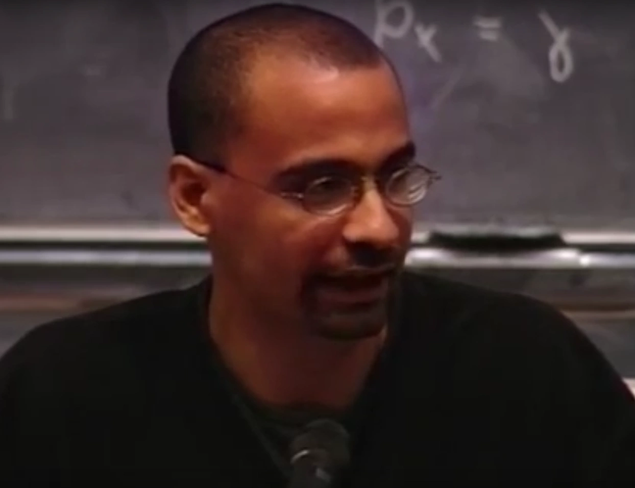 Junot Diaz looks to the side, seated in front of a chalkboard