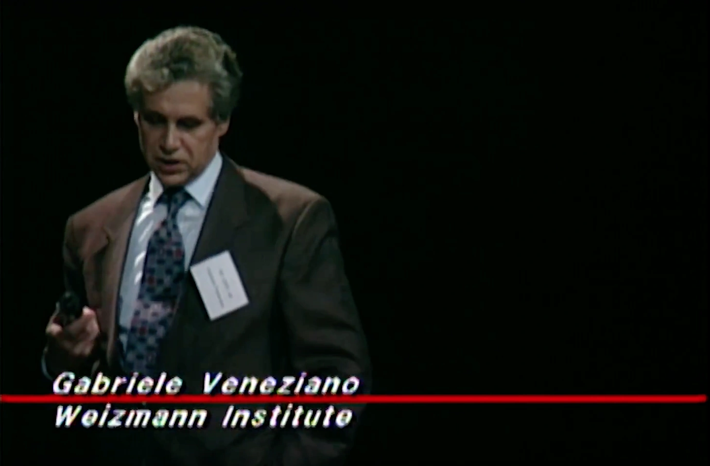 Gabriele Veneziano speaking on stage, looking downward, in front of dark background