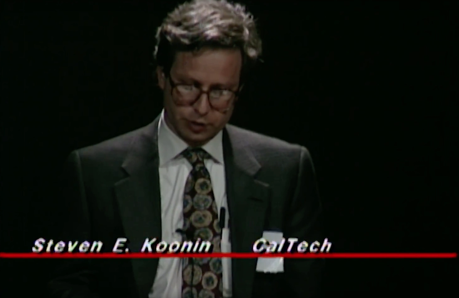 Steven E. Koonin speaking at a podium on stage, in front of a dark background