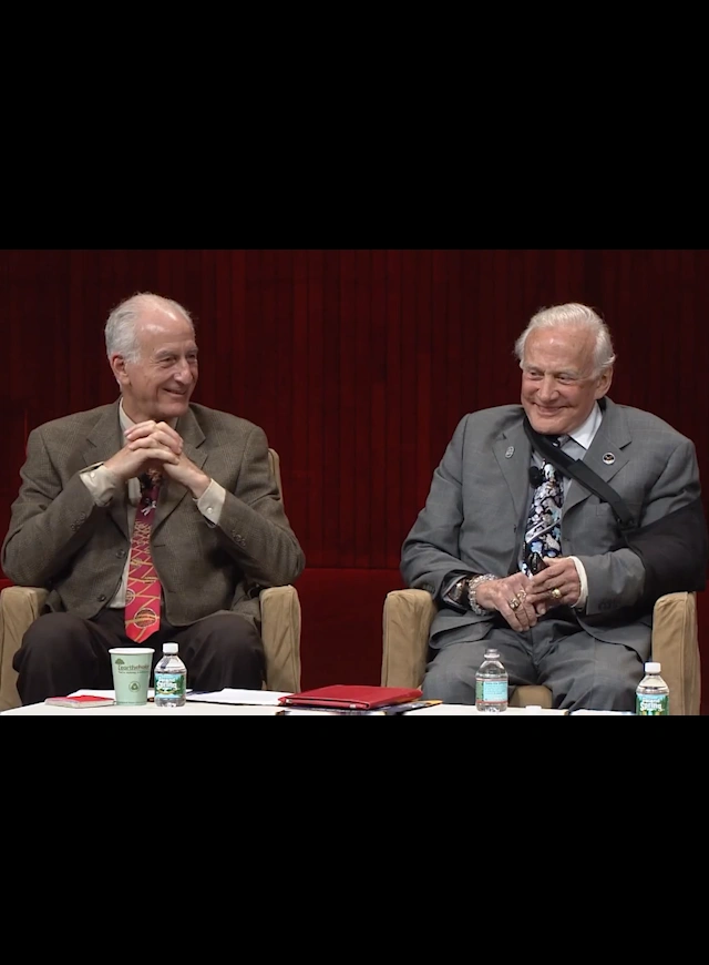 A panel of MIT alumni are seated in chairs on stage for a discussion, red curtain in background.