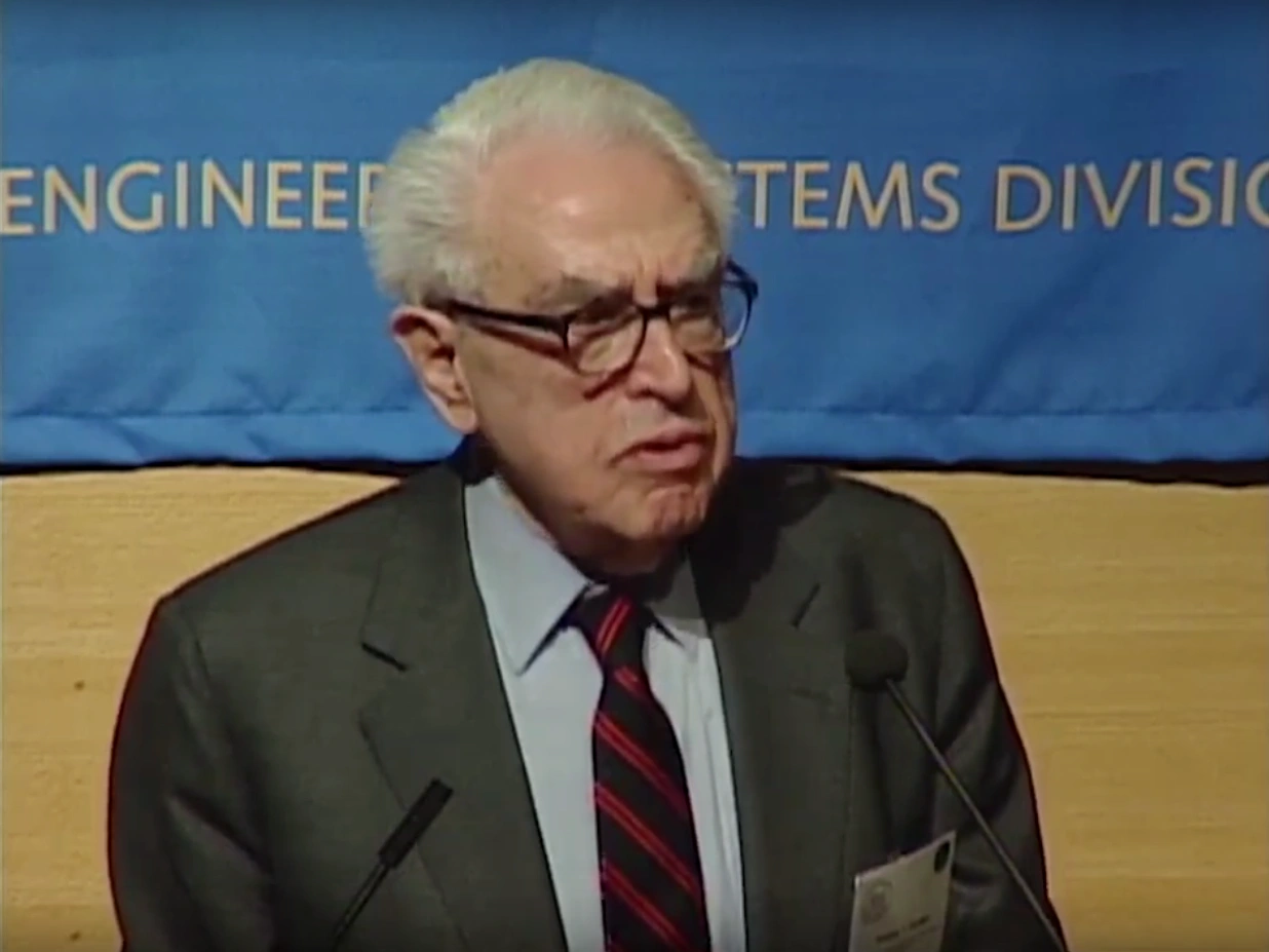 Charles M. Vest speaking at podium outside, large blue banner hanging on wood paneled wall with a in the background.