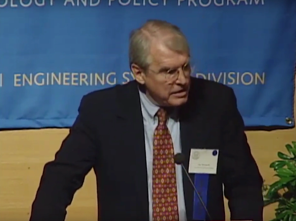 Eugene B. Skolnikoff speaking at podium outside, large blue banner hanging on wood paneled wall with a in the background.