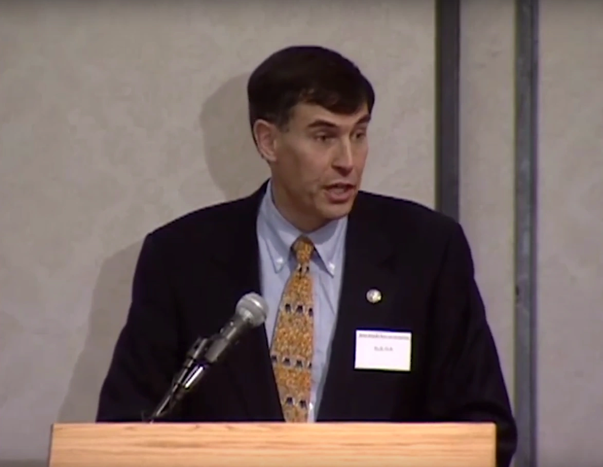 Rush Holt speaking at a podium, cream colored wall in background