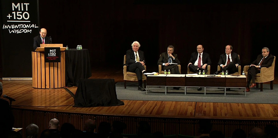One person stands behind a podium introducing panelists seated in chairs across the stage.