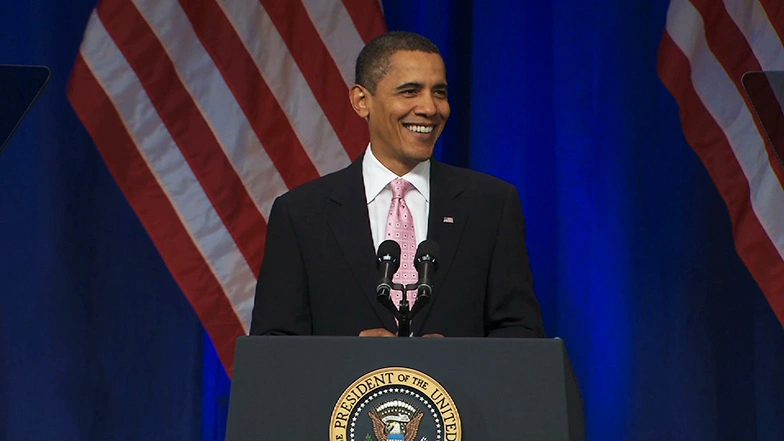 President Barack Obama smiles behind podium onstage, a blue screen and American flags in the background.