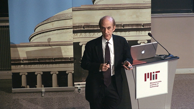 Stephen Lippard speaking at podium on stage, standing in front of backdrop picturing sections of MIT buildings.